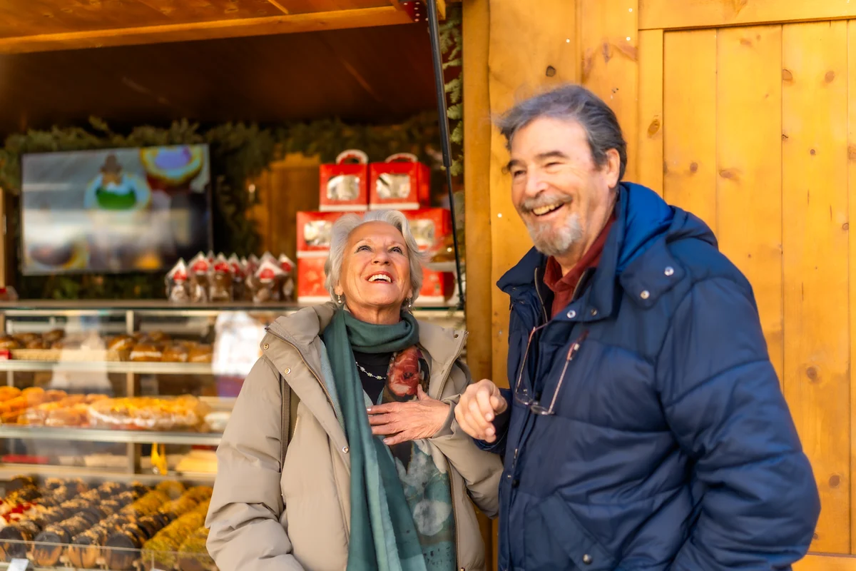 A senior couple laughing together at a Christmas market stall stacked with gingerbread and chocolates