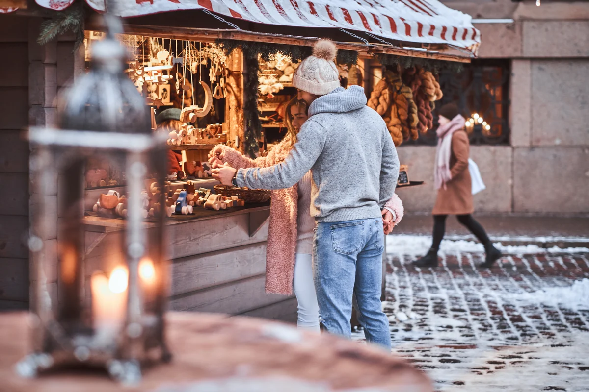 A couple browsing hand-carved wooden ornaments at a snow-dusted market stall, a warm lantern glowing in the foreground