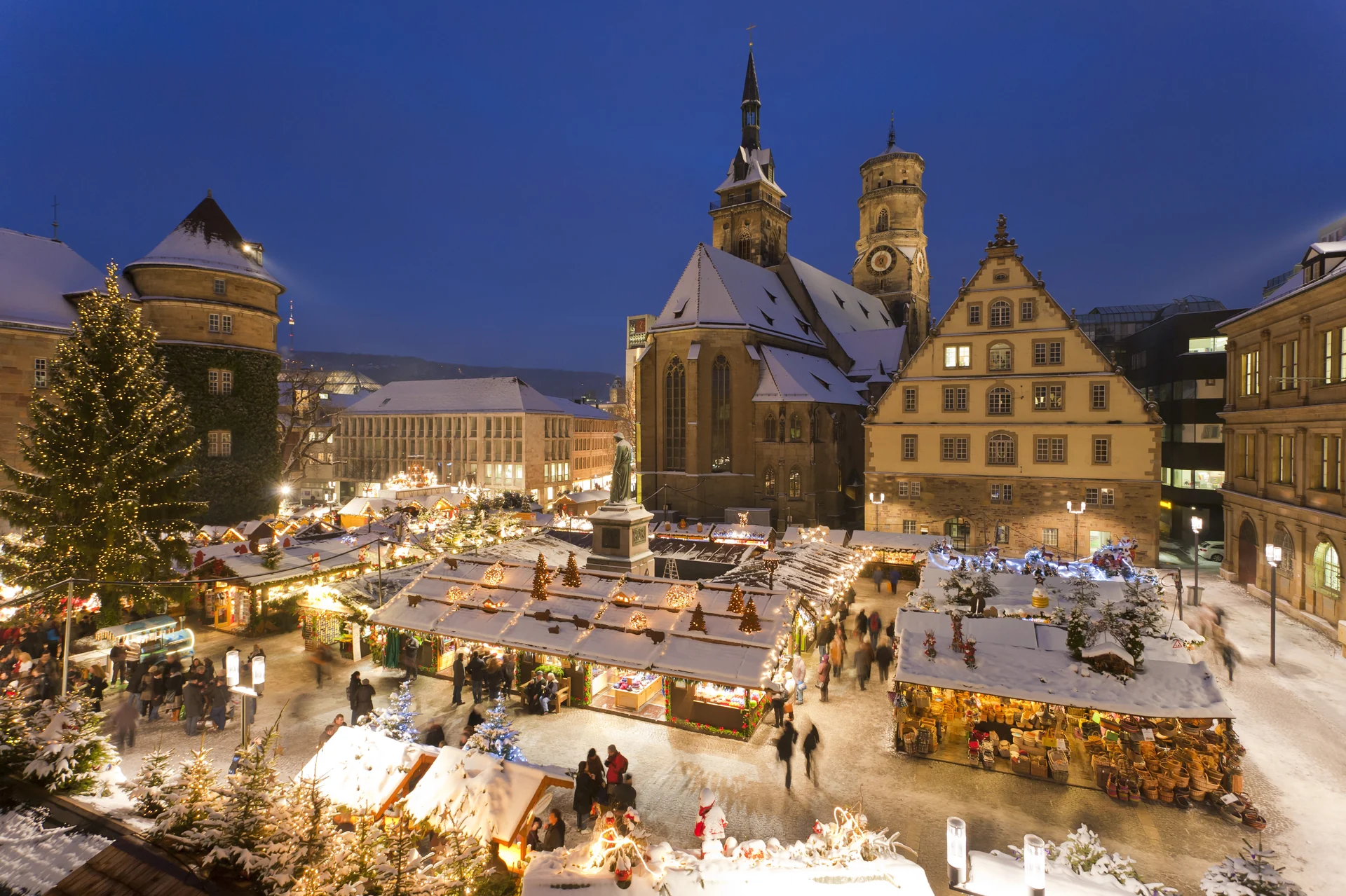 A grand European Christmas market at dusk, stalls aglow with warm light beneath illuminated church spires