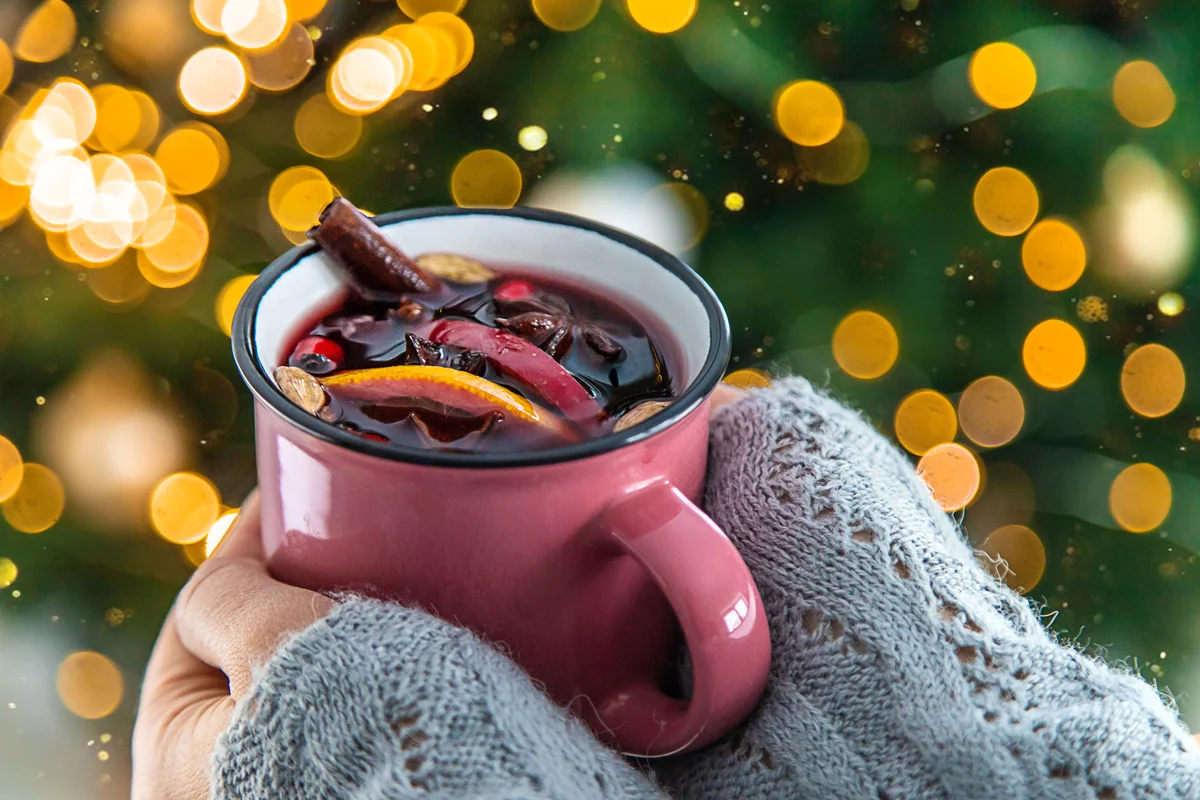 A pink ceramic mug of mulled wine with cinnamon stick, orange slice and cranberries, held by sweatered hands against a golden bokeh background