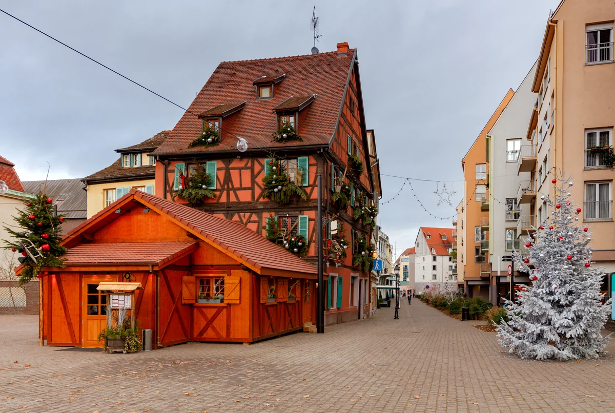 A charming European street with festive half-timbered buildings, a wooden market cabin and frosted Christmas tree