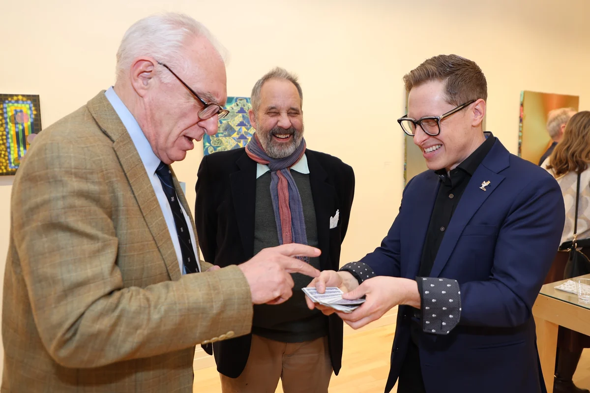 A magician laughing while sharing a card trick with two older gentlemen in an intimate gallery setting