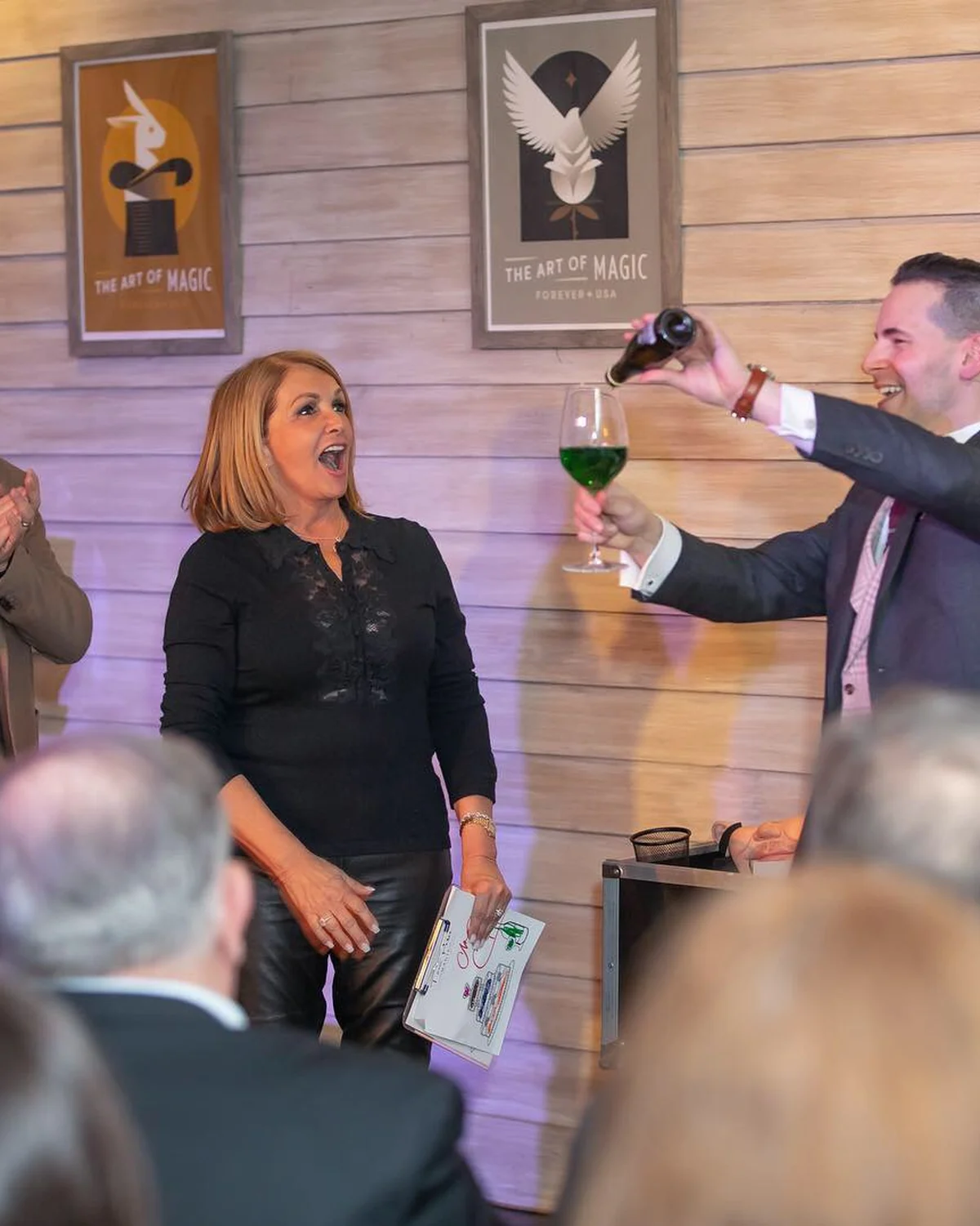 A magician pouring liquid into a wine glass that has impossibly turned green, while the guest beside him gasps in delighted disbelief