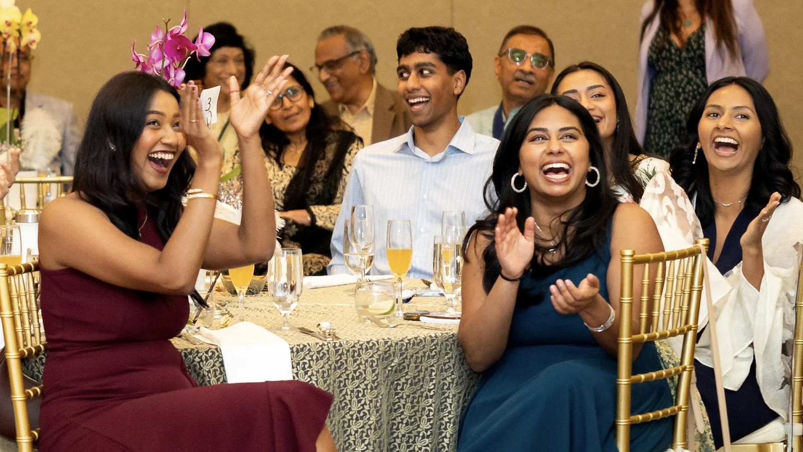 A table of elegantly-dressed guests at a formal event, faces lit up mid-laugh and applauding as a magician performs