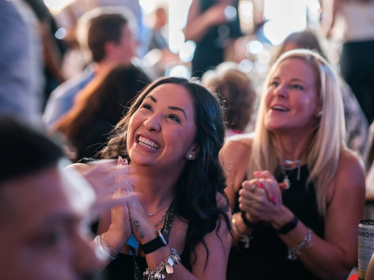 Two women in the audience laughing together, the woman in front beaming with hands clasped in delight as the woman next to her claps along