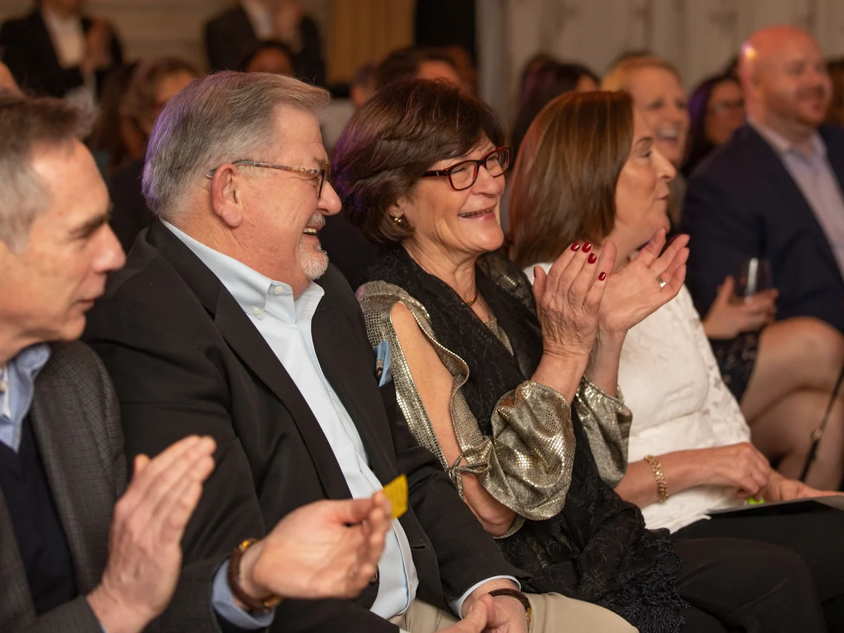 A row of seated guests laughing and clapping together, a couple in the middle catching each other's eyes as they react to the magic